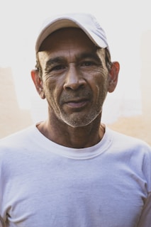A middle-aged man with short hair and a well-worn expression stands against a light, neutral background. He is wearing a white t-shirt and a white cap, suggesting a casual or outdoor setting.
