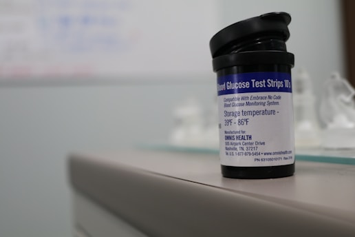 A container labeled 'Blood Glucose Test Strips' is placed on a surface. The label provides storage temperature information and details about the manufacturer. The focus is primarily on the container, with a blurred background.