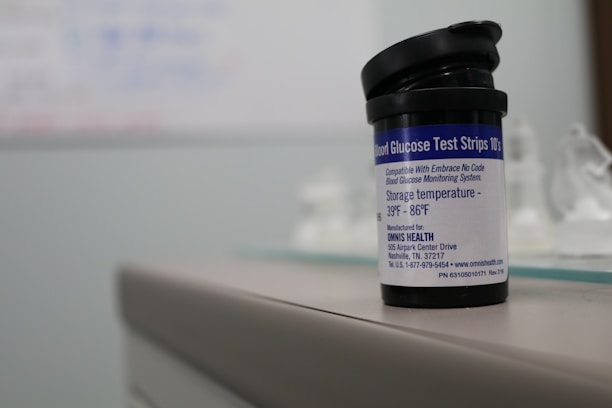 A container labeled 'Blood Glucose Test Strips' is placed on a surface. The label provides storage temperature information and details about the manufacturer. The focus is primarily on the container, with a blurred background.