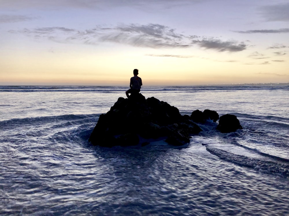 silhouette photo of person sitting on rock formation in between body of water