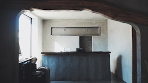 A small reception desk is set in a minimalistic room with gray concrete walls. Above the desk, there is a rectangular ventilation unit. On the left, sunlight pours in through a large window, creating soft shadows. A couple of suitcases are visible near the window, and a sign labeled 'Key Drop' sits on the desk.