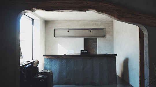 A small reception desk is set in a minimalistic room with gray concrete walls. Above the desk, there is a rectangular ventilation unit. On the left, sunlight pours in through a large window, creating soft shadows. A couple of suitcases are visible near the window, and a sign labeled 'Key Drop' sits on the desk.