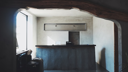 A small reception desk is set in a minimalistic room with gray concrete walls. Above the desk, there is a rectangular ventilation unit. On the left, sunlight pours in through a large window, creating soft shadows. A couple of suitcases are visible near the window, and a sign labeled 'Key Drop' sits on the desk.