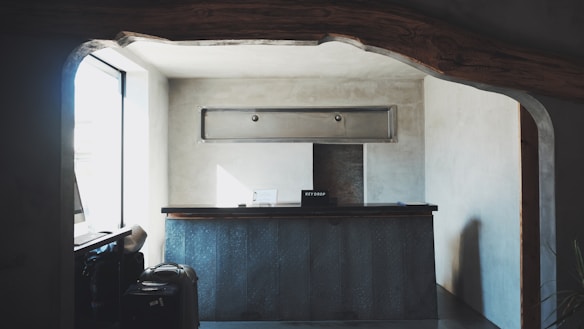 A small reception desk is set in a minimalistic room with gray concrete walls. Above the desk, there is a rectangular ventilation unit. On the left, sunlight pours in through a large window, creating soft shadows. A couple of suitcases are visible near the window, and a sign labeled 'Key Drop' sits on the desk.