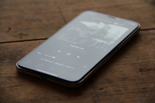A smartphone lies on a wooden table, displaying a music player interface. The screen shows a song currently playing, with visible play and pause controls, as well as a progress bar.