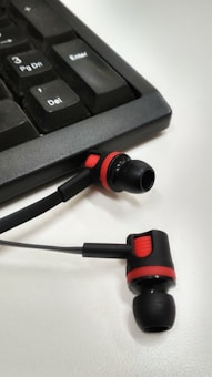 A set of black and red in-ear headphones positioned on a white surface next to a black computer keyboard. The headphones have a distinctive red ring design around the earbuds and a flat cable extending from them.