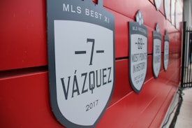A red wall displays several black and white shield-shaped plaques with labels and dates. The foreground plaque reads '-7 VÁZQUEZ 2017' and mentions 'MLS BEST XI'. Other plaques highlight records and player achievements.