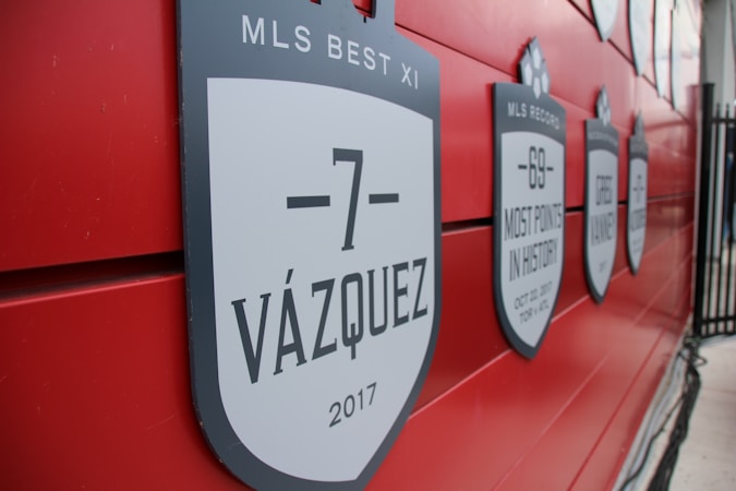 A red wall displays several black and white shield-shaped plaques with labels and dates. The foreground plaque reads '-7 VÁZQUEZ 2017' and mentions 'MLS BEST XI'. Other plaques highlight records and player achievements.