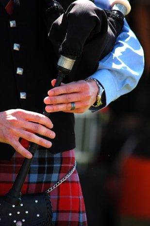 Hands are playing a set of bagpipes, with a focus on the detailed tartan kilt and black bagpipe bag. The musician is wearing a light blue shirt under a dark vest adorned with silver buttons.