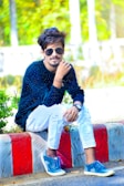 A young man with styled hair, wearing sunglasses, a dark blue long-sleeve shirt with white patterns, ripped white jeans, and blue sneakers, is sitting on a curb with a colorful red and white pattern. The background features lush green foliage and blurred elements, giving a vibrant outdoor setting.