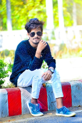 A young man with styled hair, wearing sunglasses, a dark blue long-sleeve shirt with white patterns, ripped white jeans, and blue sneakers, is sitting on a curb with a colorful red and white pattern. The background features lush green foliage and blurred elements, giving a vibrant outdoor setting.