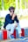 A young man with styled hair, wearing sunglasses, a dark blue long-sleeve shirt with white patterns, ripped white jeans, and blue sneakers, is sitting on a curb with a colorful red and white pattern. The background features lush green foliage and blurred elements, giving a vibrant outdoor setting.