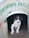 A small black and white puppy is sitting inside a wooden shelter with the words 'COROHAWK SHELTER' painted in green above the doorway. The shelter appears to be simple and rustic, with a metal rim around the entrance.