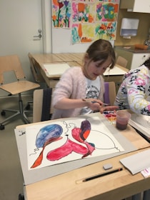 A young girl is engaged in painting with watercolors at a classroom table. Her artwork features bright, abstract shapes in colors like red, blue, and purple. Art supplies, including a paint set, a cup with water, and a pencil, are visible on the table. The background displays colorful children's art on the wall.