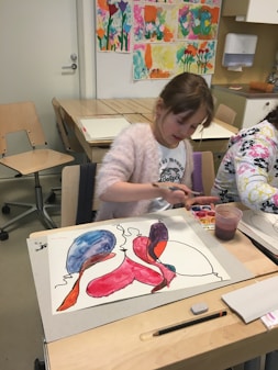 A young girl is engaged in painting with watercolors at a classroom table. Her artwork features bright, abstract shapes in colors like red, blue, and purple. Art supplies, including a paint set, a cup with water, and a pencil, are visible on the table. The background displays colorful children's art on the wall.