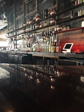 A well-stocked bar with a variety of liquor bottles displayed on shelves against a dark wooden backdrop. In the foreground, there is a shiny surface reflecting the bottles, and a small chalkboard with writing is visible among the bottles. Copper or bronze decorative elements, along with a cash register or point of sale system, are also present.