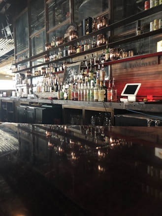 A well-stocked bar with a variety of liquor bottles displayed on shelves against a dark wooden backdrop. In the foreground, there is a shiny surface reflecting the bottles, and a small chalkboard with writing is visible among the bottles. Copper or bronze decorative elements, along with a cash register or point of sale system, are also present.