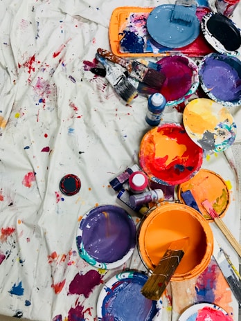 A vibrant assortment of paint cans, brushes, and paint splatters on a white canvas or cloth. The arrangement displays a range of colors, including purple, orange, blue, red, and black, along with brushes that have been used for painting. Some paint containers have paint-covered lids, and there are visible paint bottles among the scattered items.