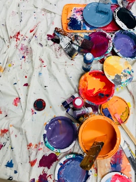 A vibrant assortment of paint cans, brushes, and paint splatters on a white canvas or cloth. The arrangement displays a range of colors, including purple, orange, blue, red, and black, along with brushes that have been used for painting. Some paint containers have paint-covered lids, and there are visible paint bottles among the scattered items.
