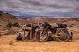 A rusty, abandoned car body is set against a vast, rugged desert landscape with distant mesas and a cloudy sky. A fully packed touring bicycle, with panniers and gear attached, leans against the car, highlighting a sense of adventure and exploration.