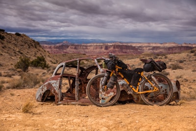 A rusty, abandoned car body is set against a vast, rugged desert landscape with distant mesas and a cloudy sky. A fully packed touring bicycle, with panniers and gear attached, leans against the car, highlighting a sense of adventure and exploration.