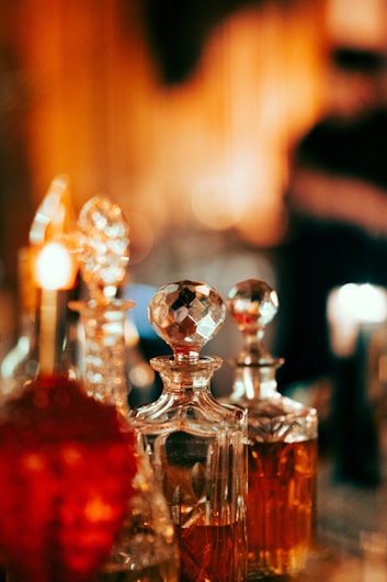 A collection of glass decanters with intricate designs and stoppers, filled with amber liquid. The setting has a warm, ambient lighting that contributes to a cozy and elegant atmosphere.
