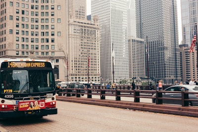 A busy urban street scene with a public bus labeled '146 Michigan Express' approaching. The background is filled with tall skyscrapers, typical of a city skyline. People are seen walking on the sidewalk and cars are driving by. The American flags add a patriotic element to the urban environment.