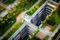 Aerial view of a modern urban landscape with buildings featuring rooftop gardens. The design incorporates green spaces and pathways, with trees and shrubs integrated into the architecture. A person walks along a path next to a structure with multiple levels.