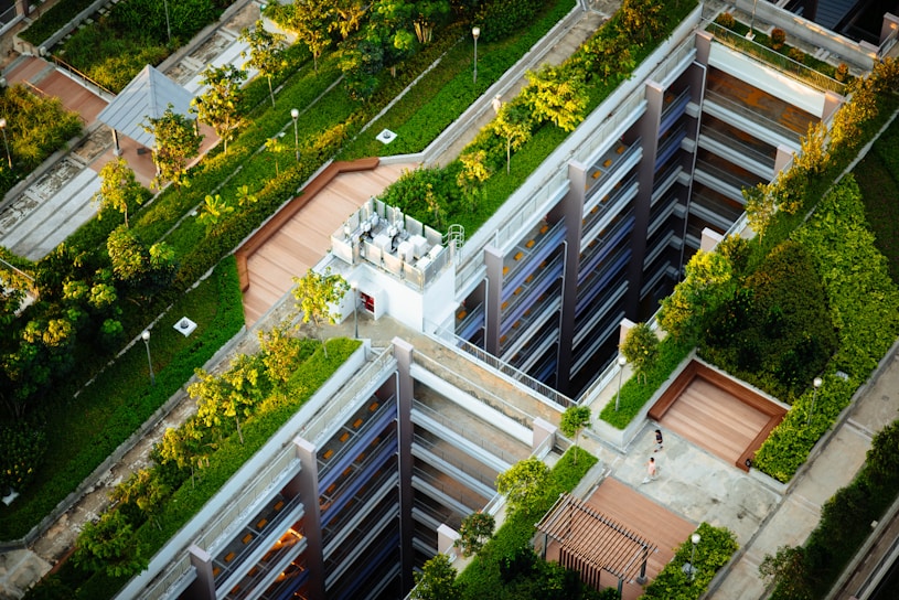 Aerial view of a modern urban landscape with buildings featuring rooftop gardens. The design incorporates green spaces and pathways, with trees and shrubs integrated into the architecture. A person walks along a path next to a structure with multiple levels.