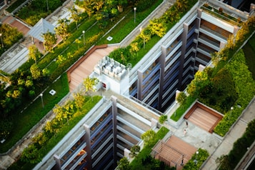 Aerial view of a modern urban landscape with buildings featuring rooftop gardens. The design incorporates green spaces and pathways, with trees and shrubs integrated into the architecture. A person walks along a path next to a structure with multiple levels.