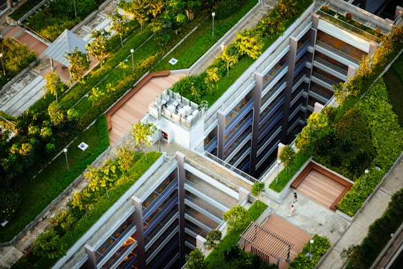 Aerial view of a modern urban landscape with buildings featuring rooftop gardens. The design incorporates green spaces and pathways, with trees and shrubs integrated into the architecture. A person walks along a path next to a structure with multiple levels.