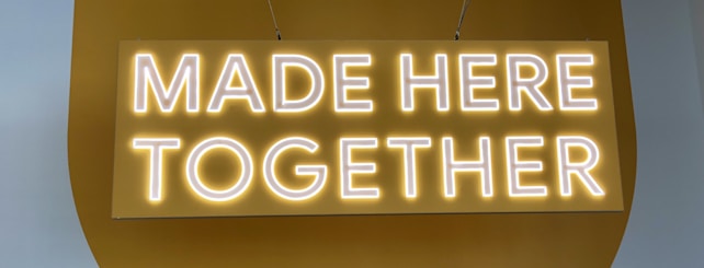 A neon sign with the words 'MADE HERE TOGETHER' displayed against a mustard-yellow background. The text is illuminated, emitting a warm, glowing light.