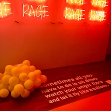A wall with multiple red neon signs displaying the word 'RAGE'. Below, a cluster of yellow balloons is placed on a wooden floor. The floor has a white text that reads: 'Sometimes all you have to do is sit down watch your anger flare and let it fly like a balloon.' The lighting creates a warm, intense ambiance.
