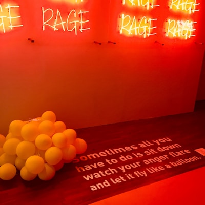 A wall with multiple red neon signs displaying the word 'RAGE'. Below, a cluster of yellow balloons is placed on a wooden floor. The floor has a white text that reads: 'Sometimes all you have to do is sit down watch your anger flare and let it fly like a balloon.' The lighting creates a warm, intense ambiance.