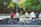 A group of youths sits on a ledge, watching a skate park where people are actively skateboarding. Their bikes are lined up in front of them, and graffiti is visible on the wall they're sitting on. The scene is set in a park with lush green trees and some buildings in the background.