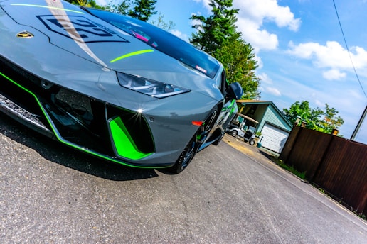 A sleek gray sports car with neon green accents parked on a paved driveway. In the background, there are trees, a small building, and some parked golf carts. The car's sharp angles and low profile emphasize its high-performance design.