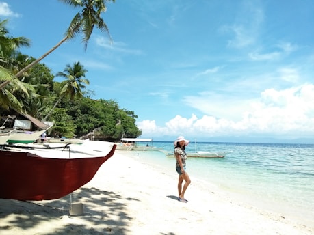 A tropical beach scene with clear blue skies and turquoise waters. A woman in a summer dress and hat stands on the white sandy shore near several outrigger boats. Palm trees line the shore, providing shade and creating a serene, picturesque setting.