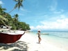 A tropical beach scene with clear blue skies and turquoise waters. A woman in a summer dress and hat stands on the white sandy shore near several outrigger boats. Palm trees line the shore, providing shade and creating a serene, picturesque setting.