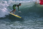 A surfer in a green and black wetsuit rides a yellow surfboard on a large wave. The waves are vibrant and turbulent, with white foam at the crest. The lighting suggests a sunny day with clear ocean water.