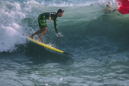 A surfer in a green and black wetsuit rides a yellow surfboard on a large wave. The waves are vibrant and turbulent, with white foam at the crest. The lighting suggests a sunny day with clear ocean water.