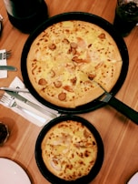Two freshly baked pizzas are placed in black pans on a wooden table. The larger pizza has toppings such as sliced sausage and cheese, while the smaller one also contains cheese. Nearby, there are cutlery and napkins, a drink in a glass, and a part of a plate can be seen.