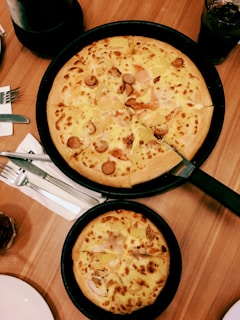 Two freshly baked pizzas are placed in black pans on a wooden table. The larger pizza has toppings such as sliced sausage and cheese, while the smaller one also contains cheese. Nearby, there are cutlery and napkins, a drink in a glass, and a part of a plate can be seen.