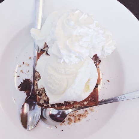 A delicious dessert with a scoop of vanilla ice cream and a generous serving of whipped cream on top. Crumbled nuts and chocolate sauce are scattered around, accompanied by two metal spoons on a white plate.