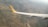 An aerial view of a landscape featuring a patchwork of fields and hills, with a close-up of an airplane wing showing a yellow tail fin with a logo. The terrain appears to be rural with a combination of green and brown patches, indicating cultivated land and natural vegetation.