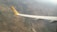 An aerial view of a landscape featuring a patchwork of fields and hills, with a close-up of an airplane wing showing a yellow tail fin with a logo. The terrain appears to be rural with a combination of green and brown patches, indicating cultivated land and natural vegetation.