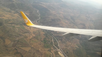 An aerial view of a landscape featuring a patchwork of fields and hills, with a close-up of an airplane wing showing a yellow tail fin with a logo. The terrain appears to be rural with a combination of green and brown patches, indicating cultivated land and natural vegetation.