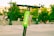 An electric scooter with a bright green label is parked outdoors. Several people are walking and sitting in the background, surrounded by trees and urban fixtures.
