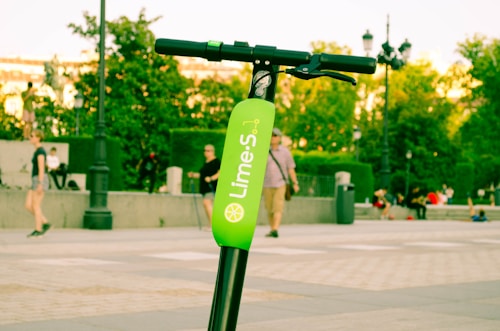 An electric scooter with a bright green label is parked outdoors. Several people are walking and sitting in the background, surrounded by trees and urban fixtures.