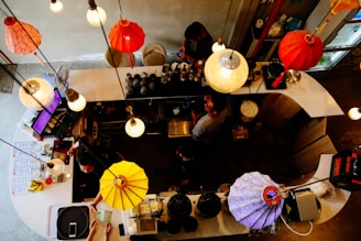Overhead view of a bustling cafe or bar counter with several illuminated hanging lamps in varying colors. Staff members are actively preparing drinks behind the counter surrounded by many bottles and equipment. Customers are handing over payment, and the area has a lively, cozy atmosphere.