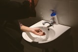 A person washing their hands under a running faucet in a sink. A liquid soap dispenser is positioned nearby on the sink's edge. Water is visibly splashing as it flows over the hands.